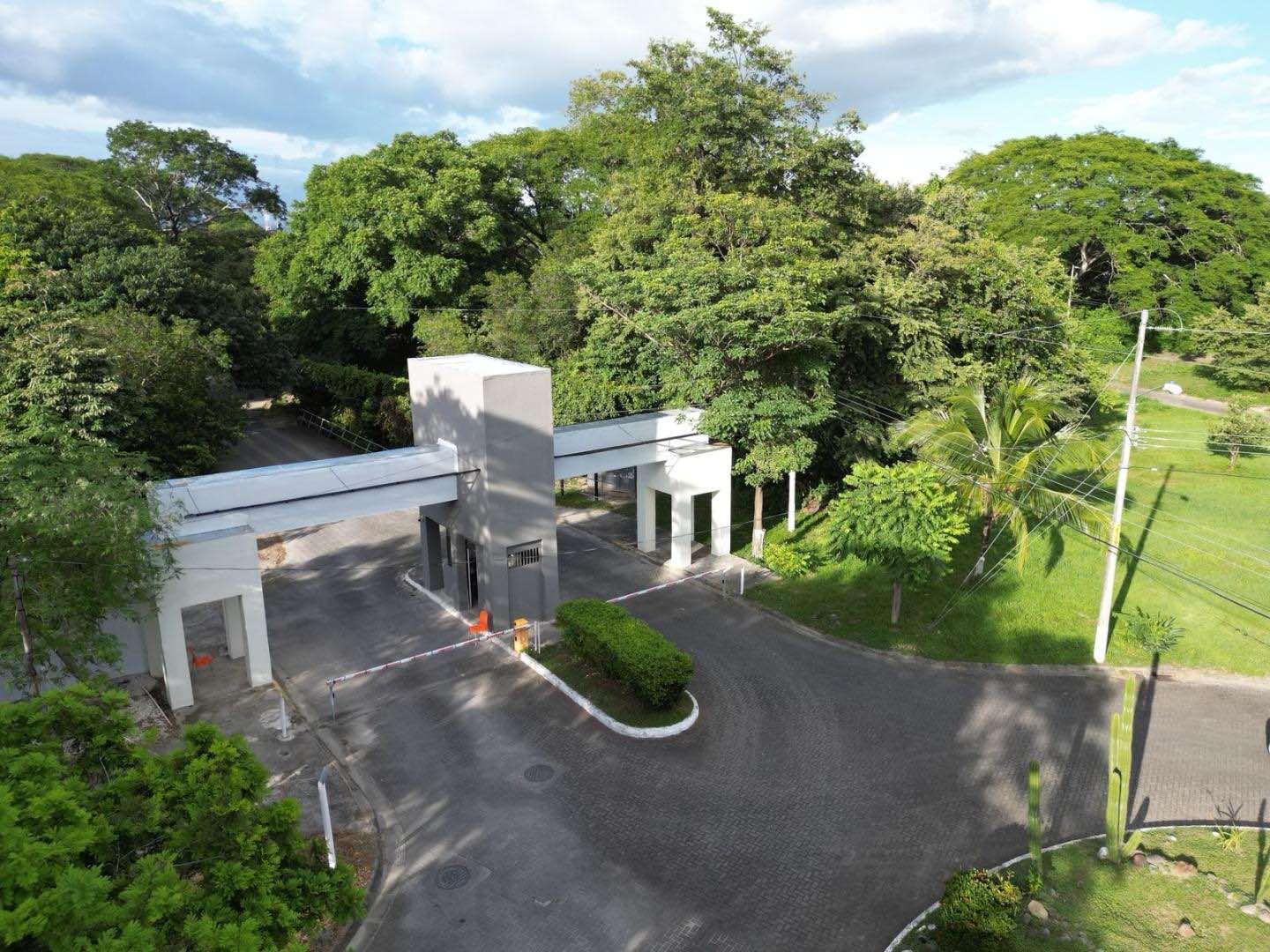 Vista aérea del pórtico de entrada de El Sitio Condominio, con la caseta de control rodeada de árboles grandes
